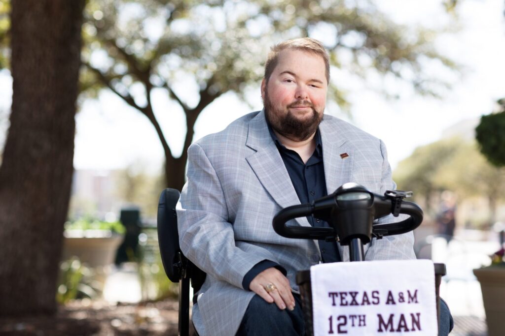 Daniel Singletary, Ed.D., seated on his mobility scooter outdoors on the Texas A&M campus, wearing a light gray blazer and his Aggie Ring, with a Texas A&M 12th Man banner draped over the handlebars.